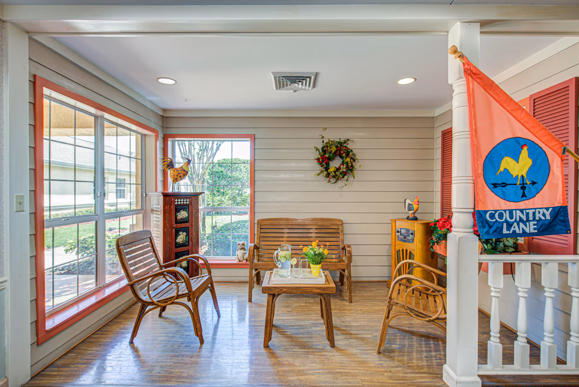 A bright and cozy sitting area with wooden chairs and a bench around a small wooden table. The space has large windows with red trim letting in natural light, a decorative wreath on the wall, and a colorful flag with a rooster and the words 'Country Lane' hanging on a white post. There are also decorative items including a rooster figurine and a small cabinet.