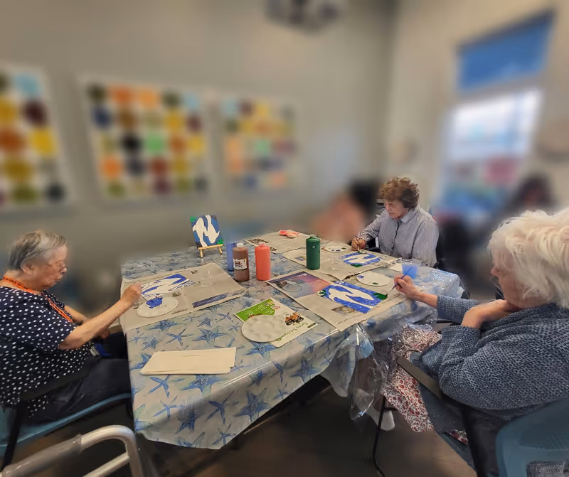 Four elderly women seated around a table covered with a starfish-patterned tablecloth, engaged in painting on canvases with blue and white colors. The room has blurred colorful artwork on the walls and a window letting in natural light.