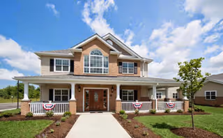 Front exterior view of a two-story residential building with a brick and siding facade, a large central window above the entrance, a covered porch with white railings decorated with red, white, and blue bunting, and a landscaped lawn with a small tree and shrubs under a partly cloudy sky.