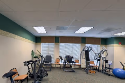 A senior living community exercise room with various fitness equipment including stationary bikes, weight machines, and a large blue exercise ball. The room has beige walls with a green accent near the ceiling, three windows with blinds, and several chairs placed around the perimeter.