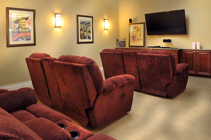 A cozy home theater room with two rows of plush red recliner chairs facing a wall-mounted flat screen TV. The walls are decorated with framed vintage movie posters and wall sconces provide warm lighting. Below the TV, there is a wooden cabinet with granite countertop holding a few items including popcorn containers.