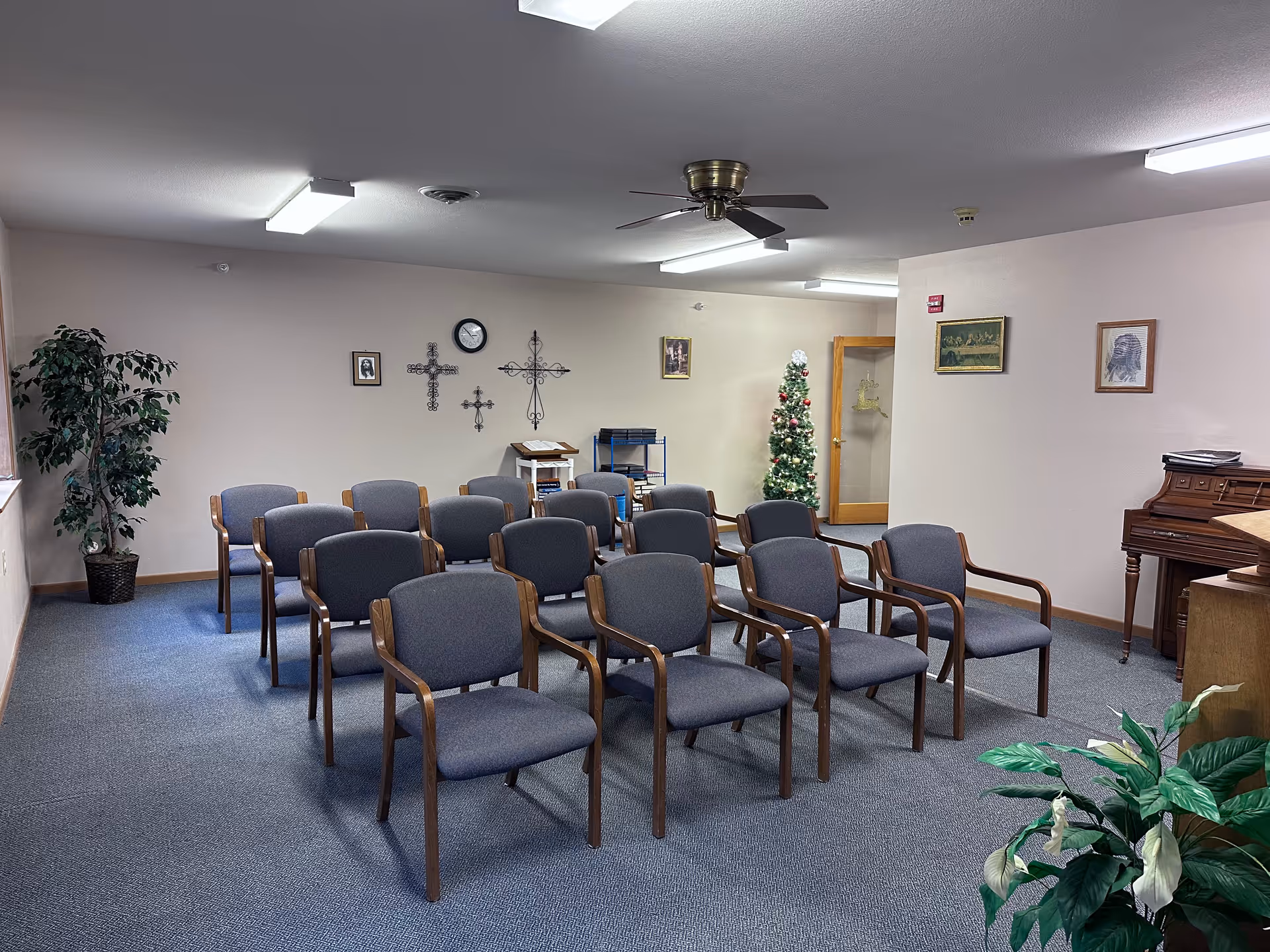 A small meeting or chapel room with three rows of gray cushioned chairs with wooden armrests arranged facing a piano and podium. The room has a ceiling fan, fluorescent lights, a decorated Christmas tree in the corner, wall decorations including crosses, framed pictures, and a clock. There are two potted plants, one near the window and one in the foreground.