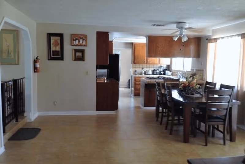 Interior view of a residential dining area and kitchen. The dining area has a dark wooden table with six matching chairs and a floral centerpiece. The kitchen features wooden cabinets, a stove, and various appliances. There is a ceiling fan above the dining table and windows with curtains allowing natural light to enter.