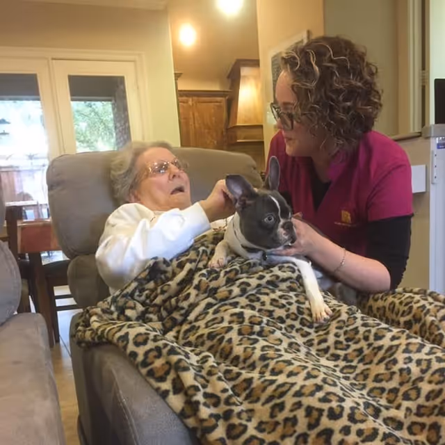 An elderly woman reclining in a comfortable chair covered with a leopard print blanket, interacting with a small black and white dog held by a caregiver wearing glasses and a magenta uniform top in a cozy living room setting.