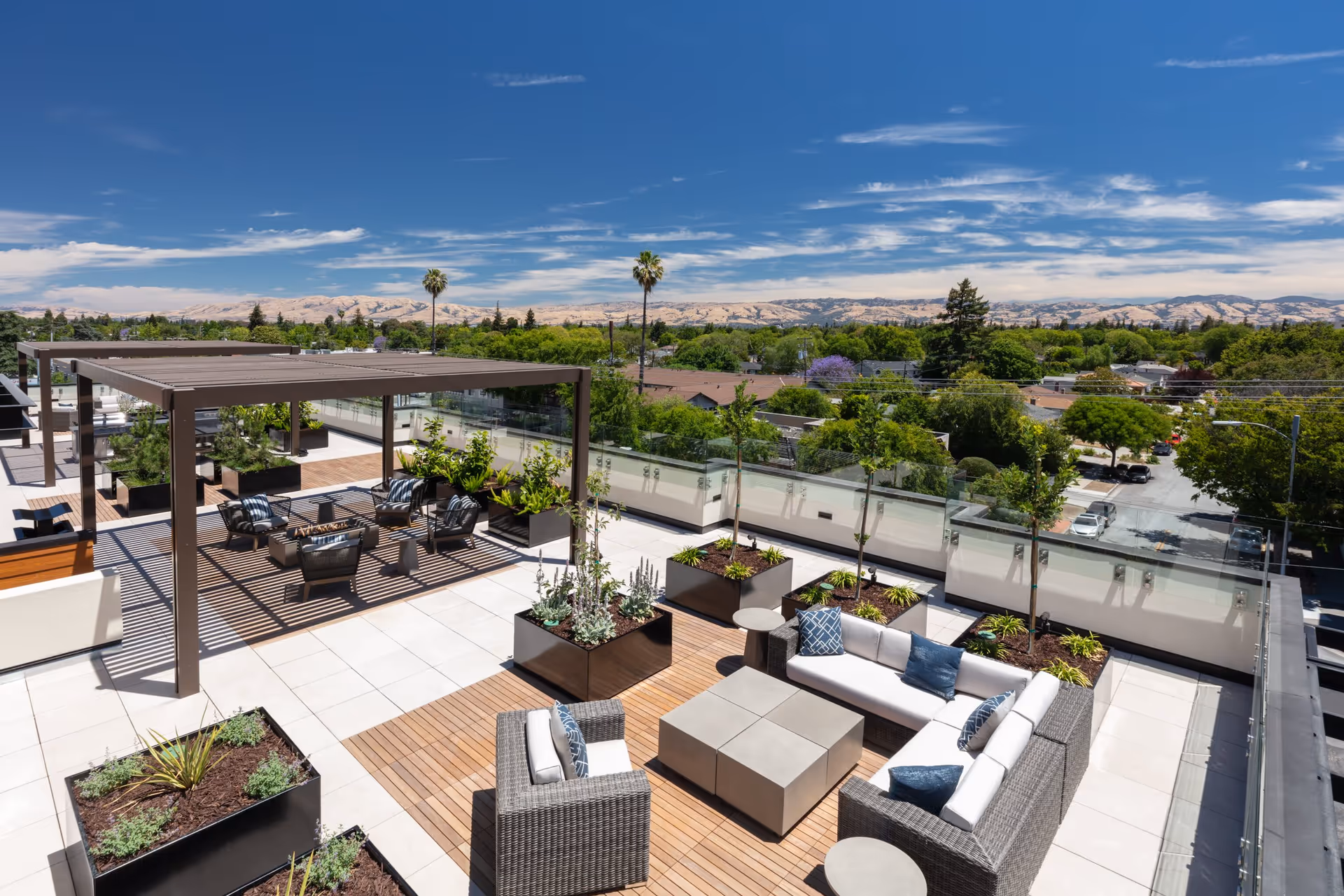 Rooftop terrace with shaded pergolas, lounge seating, planters and a panoramic view of trees and distant hills.
