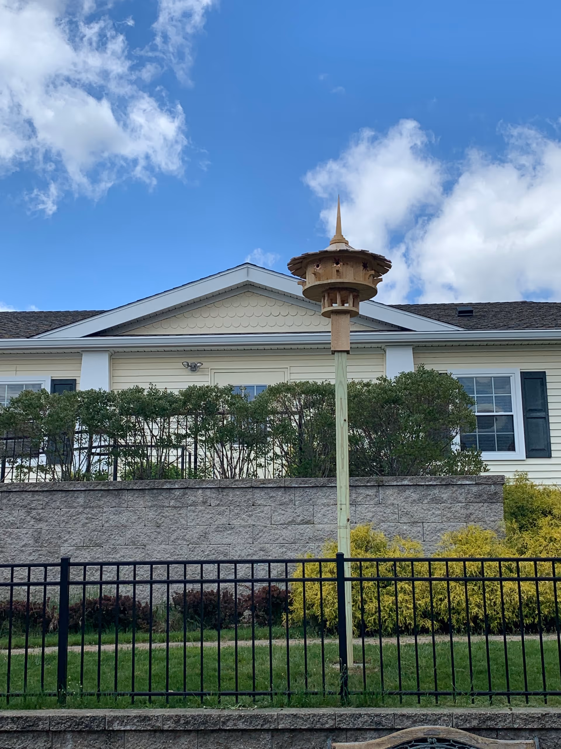 Exterior view of a single-story building with a decorative multi-tiered birdhouse on a tall post, landscaping and a black metal fence under a blue sky.