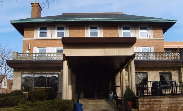Brick senior living facility with a covered entrance, pillars, and balcony on a two-story facade.