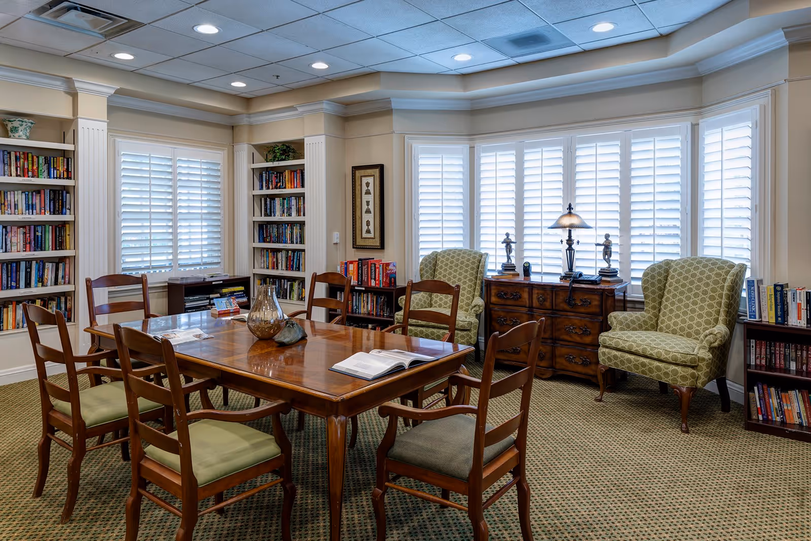 A bright community reading room with a central wooden table and chairs, bookshelves lining the walls, and upholstered armchairs by shuttered windows.
