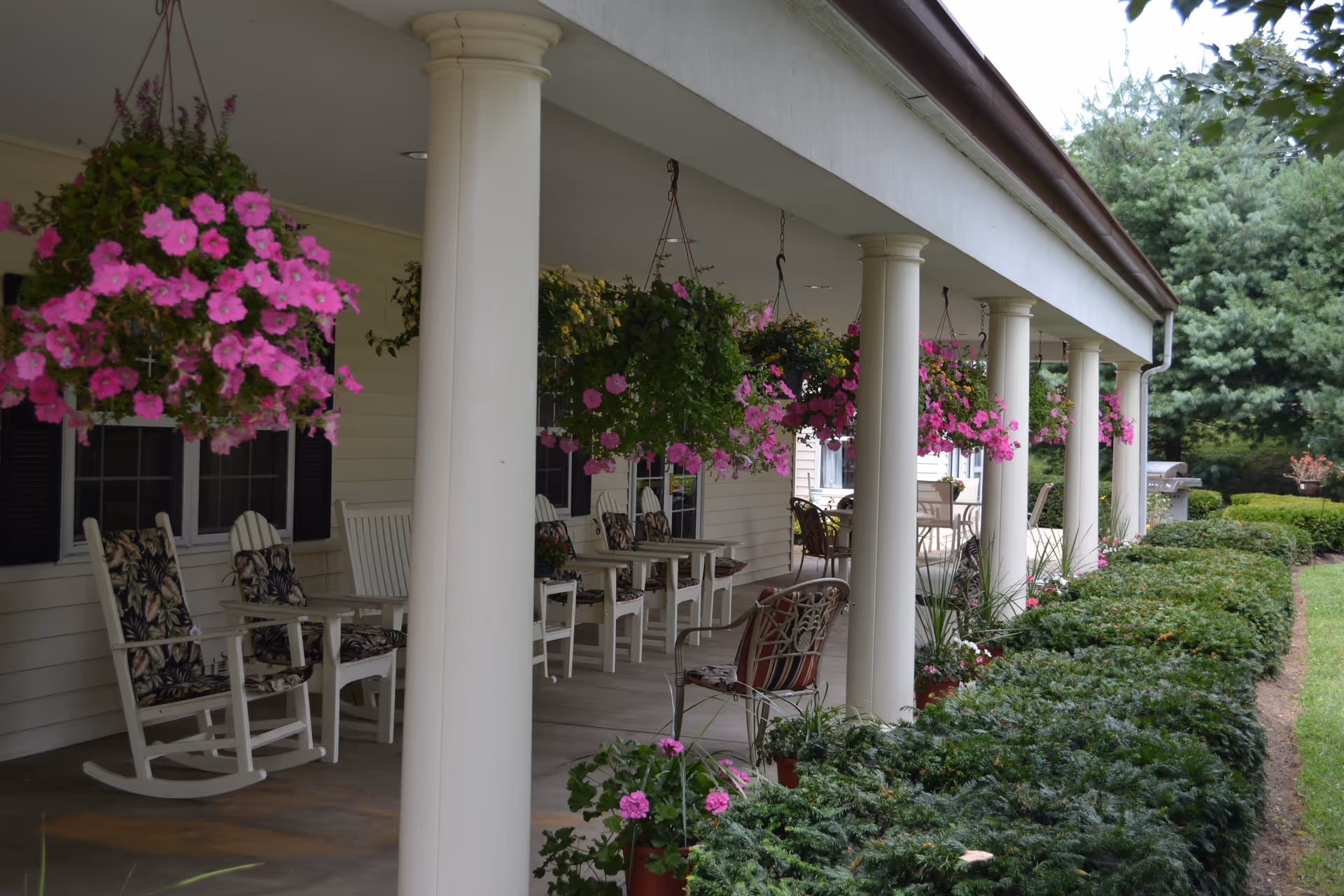Covered porch with white columns, hanging pink flower baskets, rocking chairs and seating along a senior living facility.