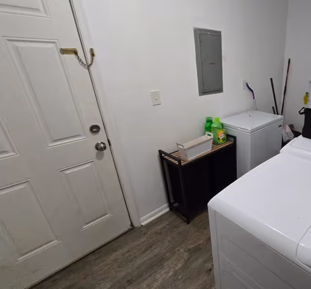 Laundry room with a white washing machine and a white chest freezer. There is a small black shelf holding laundry detergent and a white basket. A closed white door with a chain lock is on the left side, and cleaning tools are visible in the corner.