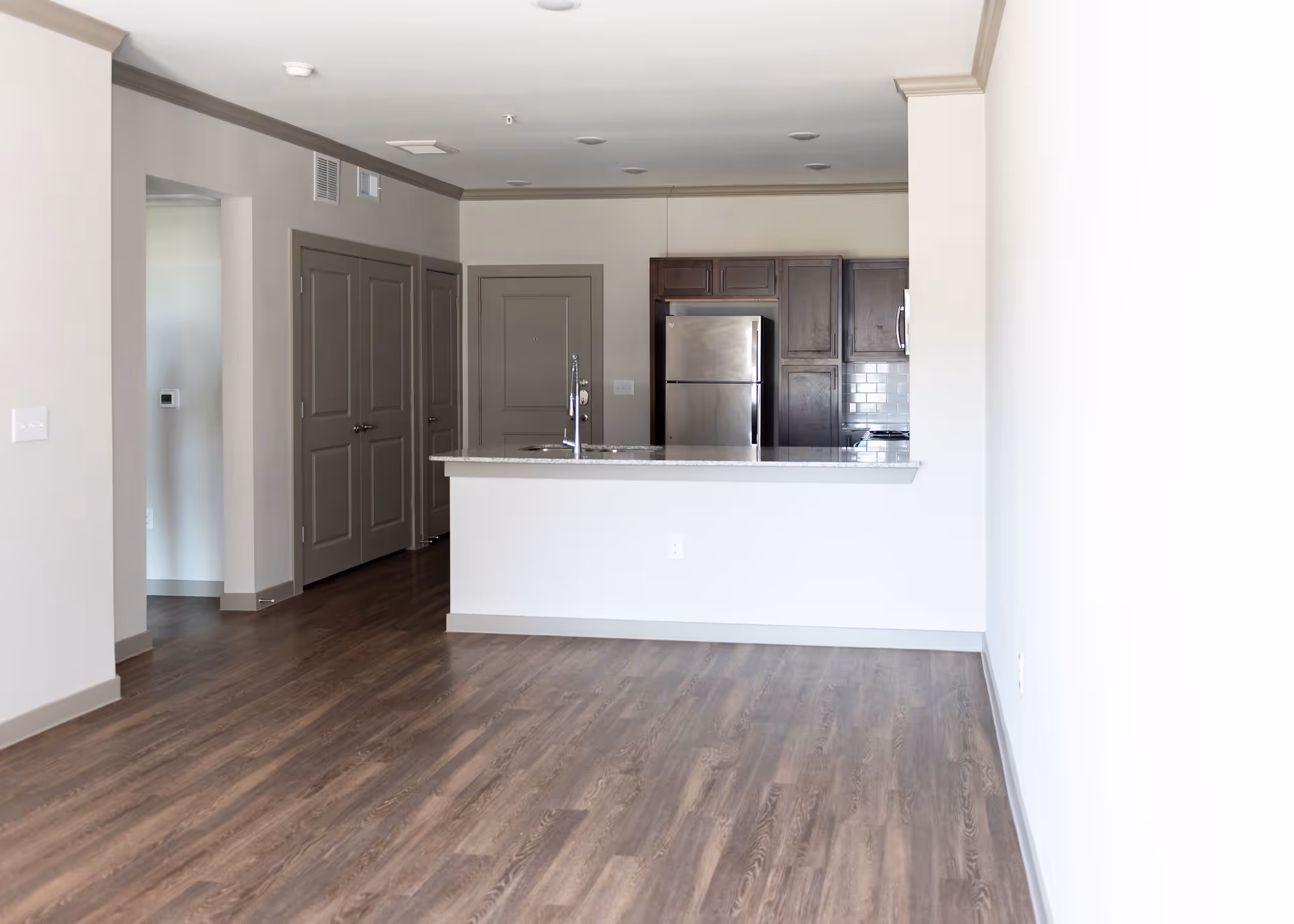 Empty modern apartment interior featuring a kitchen with dark wood cabinets, stainless steel refrigerator, and a granite countertop island with a sink. The room has light-colored walls, wood flooring, and multiple closed doors along the left wall.
