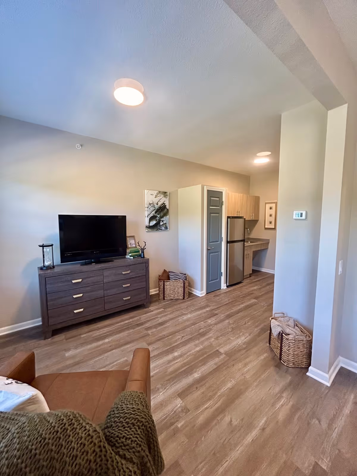 A modern living area with light wood flooring, a flat-screen TV on a dark wood dresser, a brown armchair with a green knitted throw, and a small kitchen area with a refrigerator and cabinets in the background. The walls are light-colored, and there are baskets and framed artwork decorating the space.