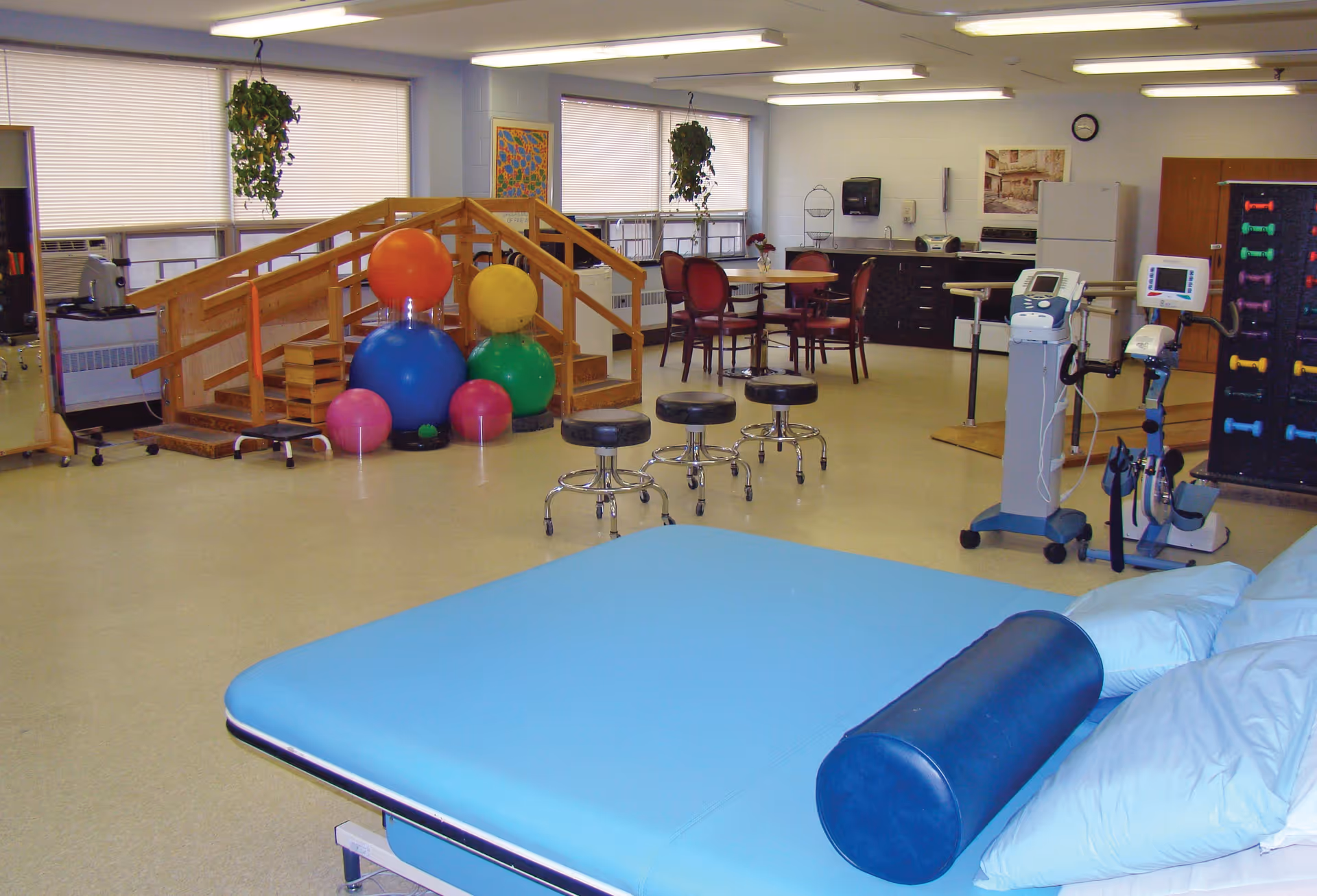 A spacious rehabilitation room with exercise equipment including colorful therapy balls, a wooden ramp with handrails, stools on wheels, and medical devices. There is a blue padded therapy table with pillows and a bolster in the foreground. In the background, there is a small dining area with a table and chairs, a kitchenette with a refrigerator, and a rack with colorful dumbbells.