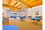 Spacious dining room with multiple tables covered in blue tablecloths, wooden chairs with blue cushions, large windows with curtains, and a wooden ceiling with exposed beams and modern chandeliers providing lighting.