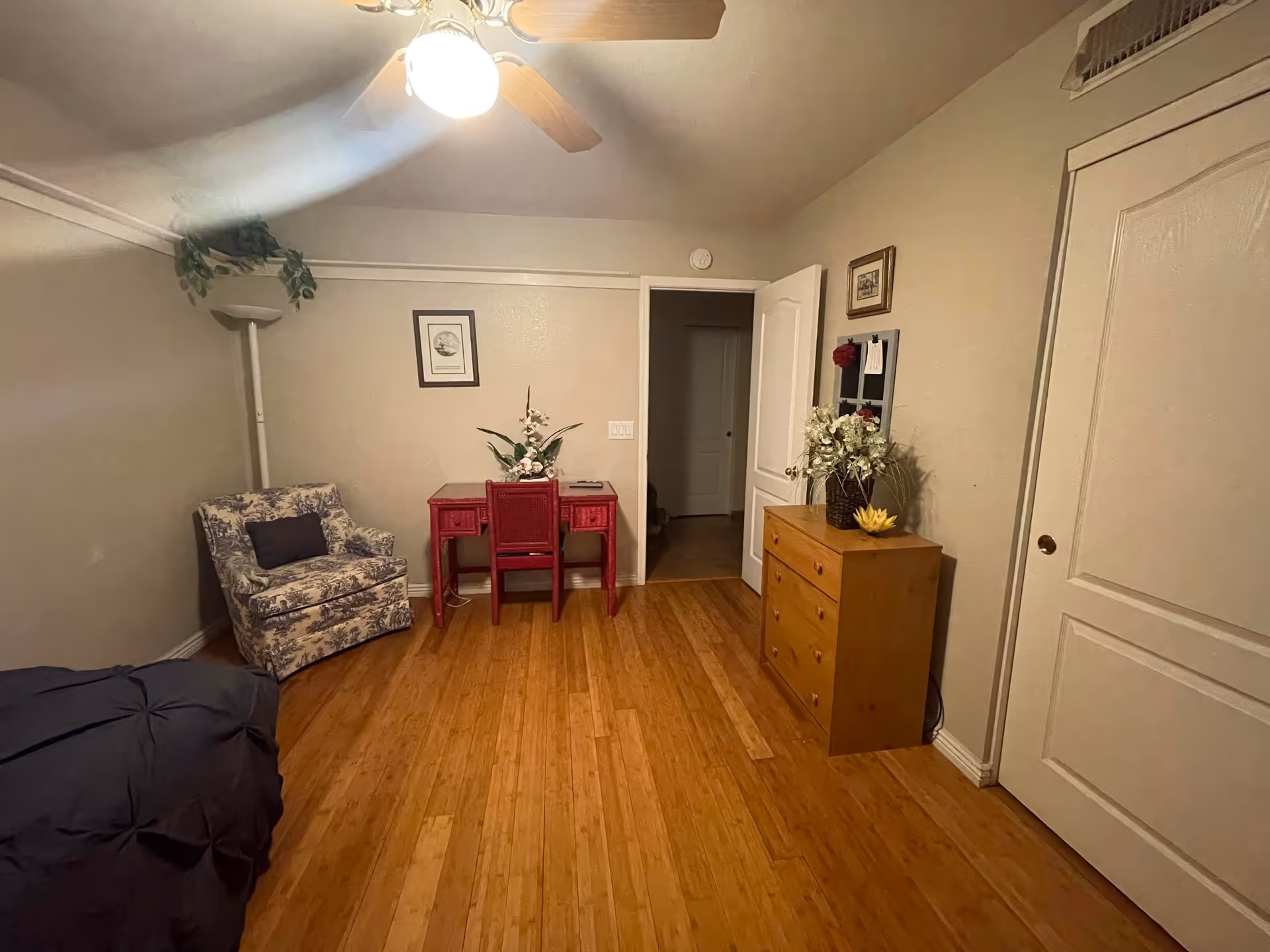 A cozy bedroom with wooden flooring, a ceiling fan with light, a floral armchair with a dark cushion, a red desk with a matching chair and a flower arrangement on top, a wooden dresser with a flower vase and decorative item, and two closed doors on the right wall. The walls are light-colored with framed pictures and a floor lamp in the corner.