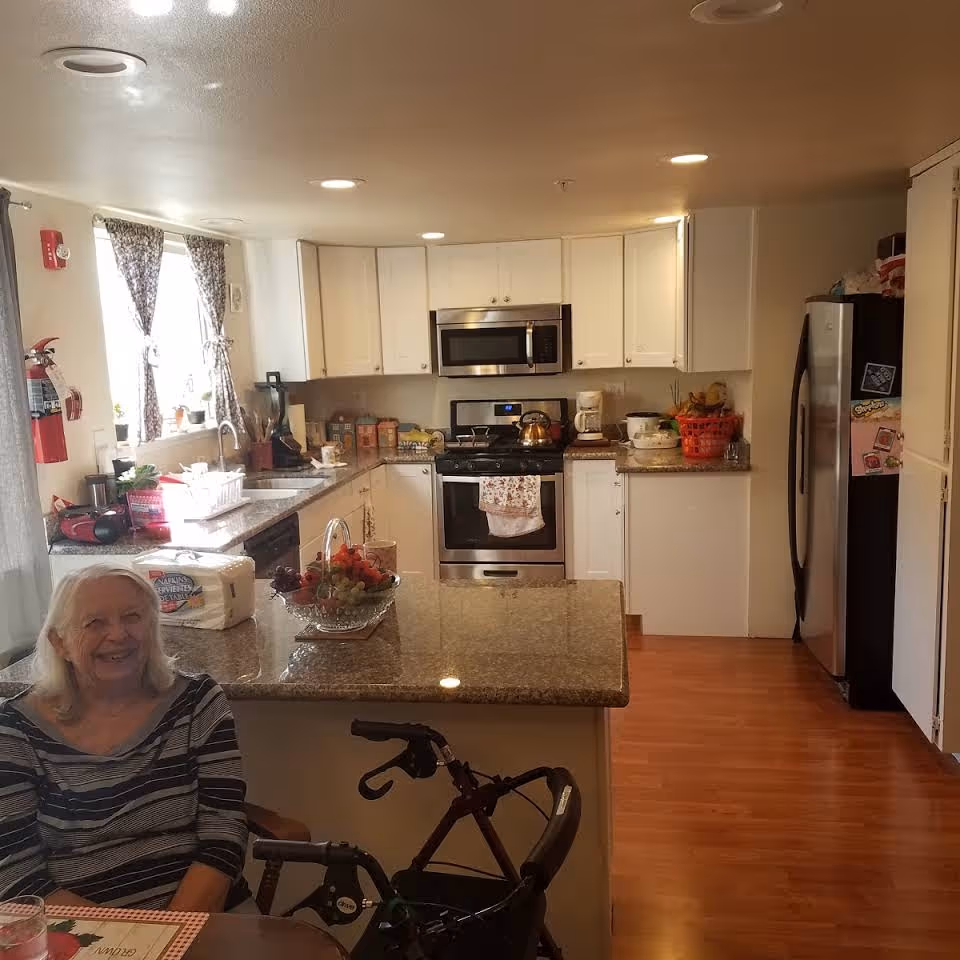 A bright kitchen with white cabinets, granite countertops, and wooden flooring. The kitchen features a stainless steel microwave, stove, and refrigerator. A bowl of fruit and a roll of paper towels are on the counter. An elderly woman with white hair is sitting at the counter, smiling, with a walker nearby. The kitchen window has patterned curtains and there are various kitchen items on the counters.