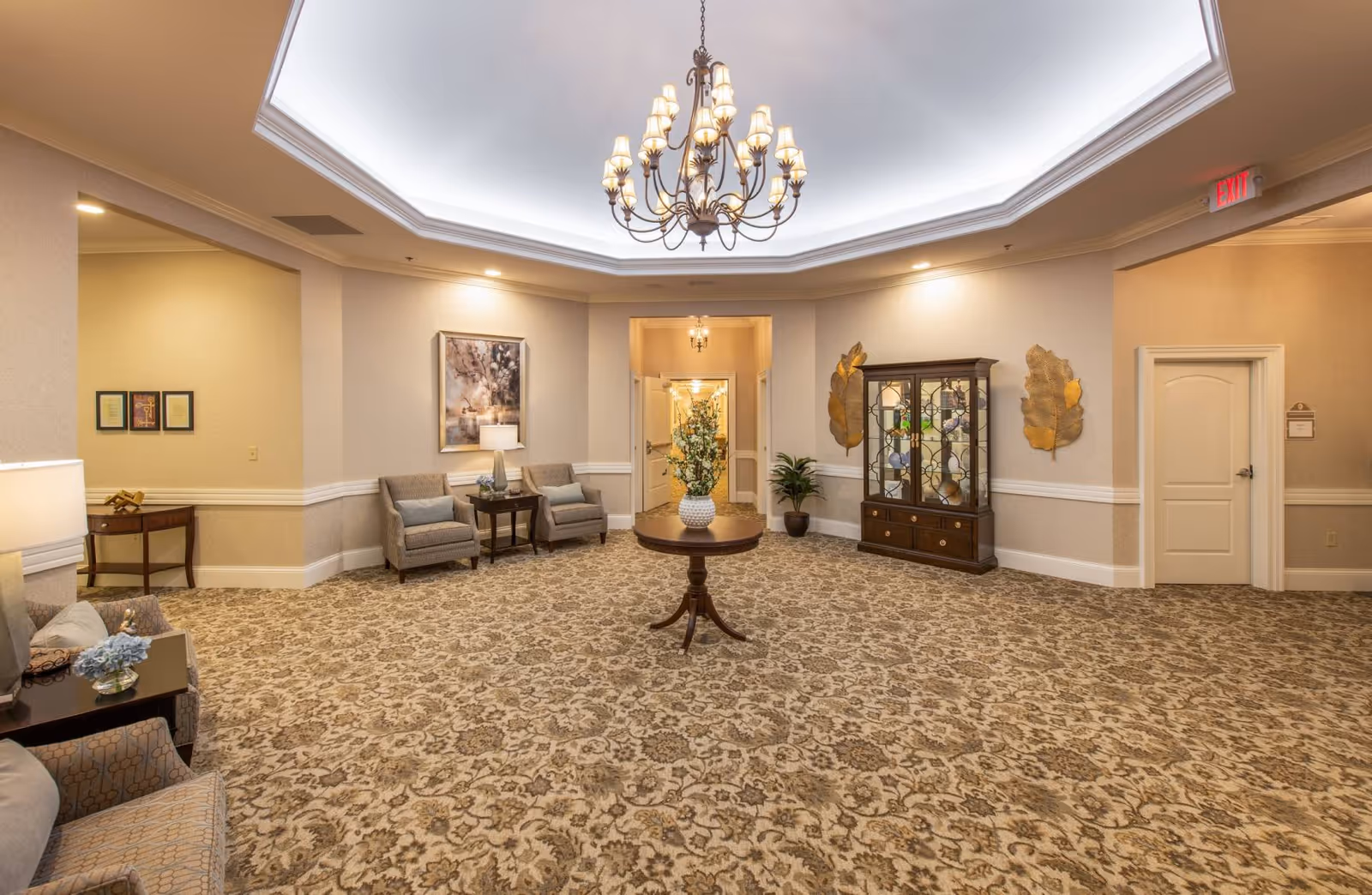 A spacious, well-lit common area with patterned carpet, beige walls, and a large chandelier hanging from a recessed ceiling. The room features a round wooden table with a vase of flowers in the center, two armchairs with a small table and lamp between them, a wooden display cabinet with decorative items, and wall art including two large gold leaf sculptures. There is an exit sign above a door on the right and an open doorway in the center leading to another room.