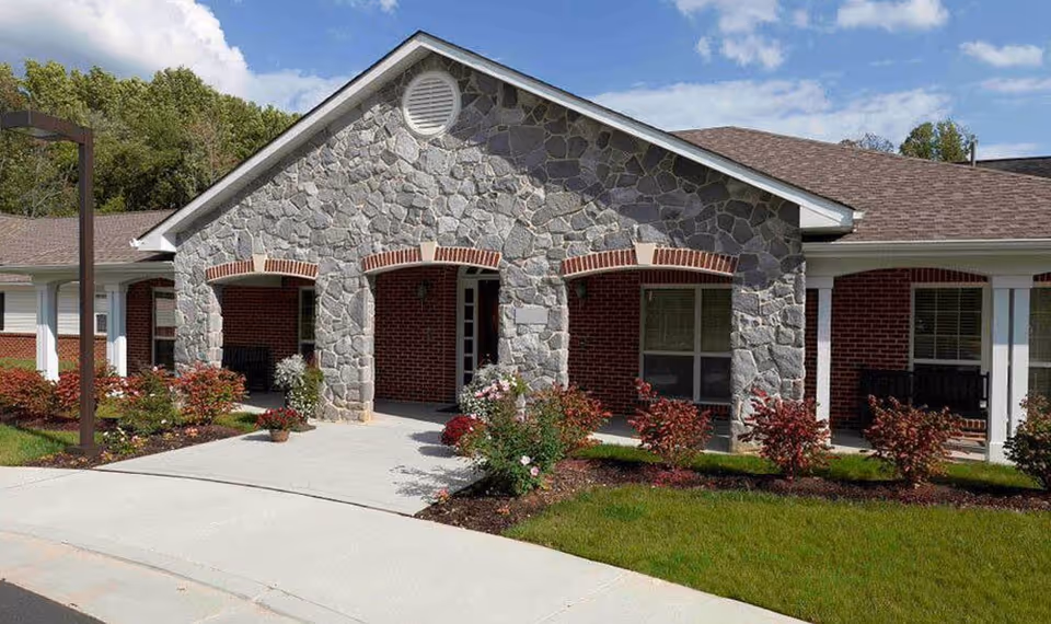 Exterior view of a senior living facility with a stone and brick facade, featuring two arched entryways, a concrete walkway, landscaped bushes, and a grassy lawn under a partly cloudy sky.