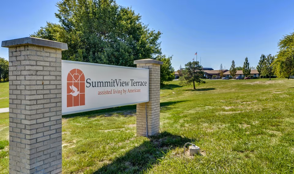 Outdoor view of SummitView Terrace assisted living facility sign mounted on two brick pillars with a grassy lawn, trees, and the facility building in the background under a clear blue sky.