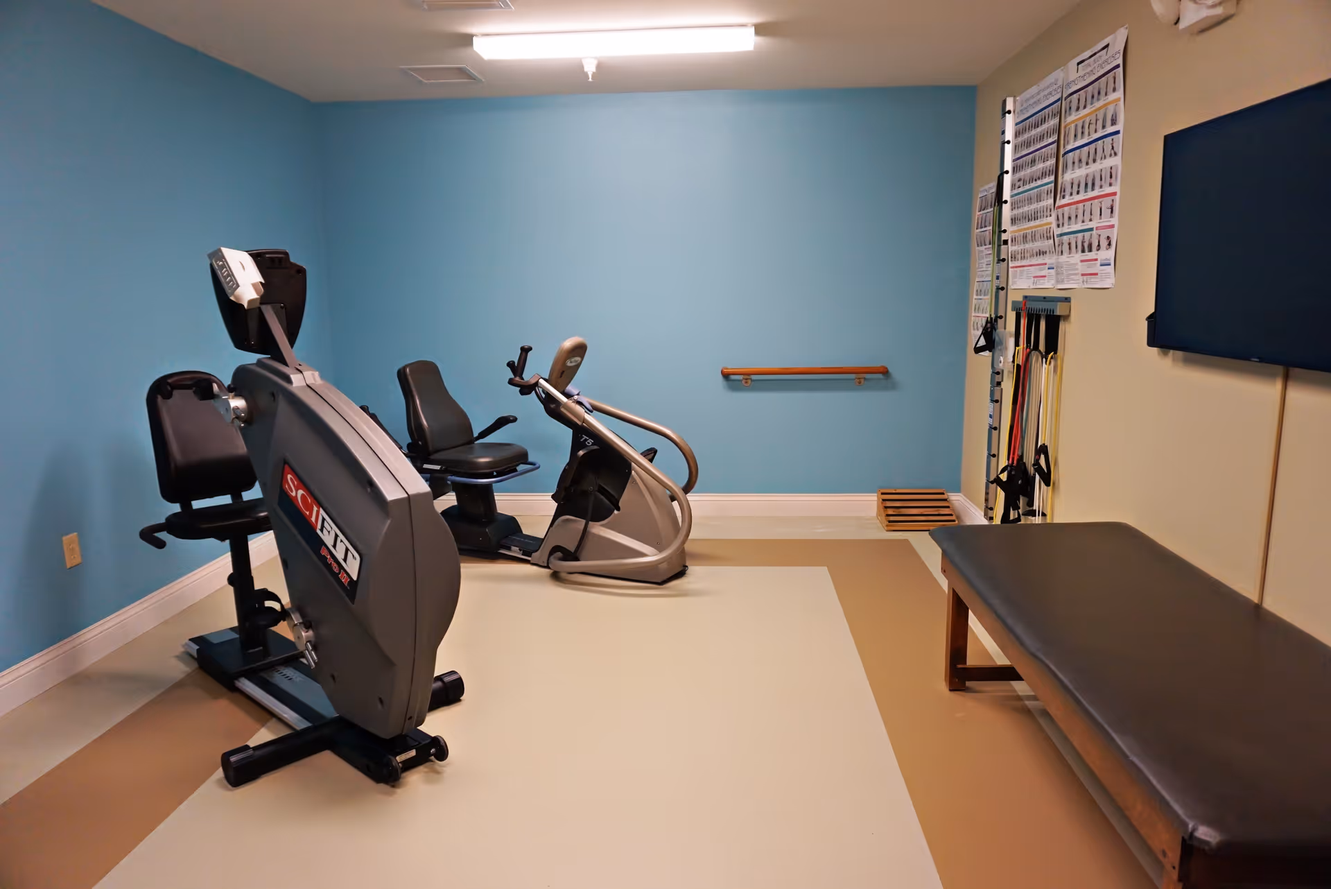 Small exercise/therapy room with two recumbent exercise bikes, a treatment table, resistance bands and instructional posters on the wall.