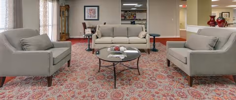 Communal seating area with three gray sofas arranged around a glass coffee table on a patterned rug and decorative accents.