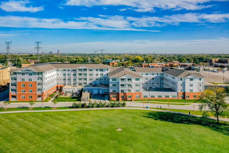 Aerial view of Clarendale of Addison, a large multi-story senior living facility with a combination of brick and light-colored siding exterior. The building is surrounded by green lawns and trees, with a clear blue sky and scattered clouds above. In the background, there are power lines and a distant cityscape.
