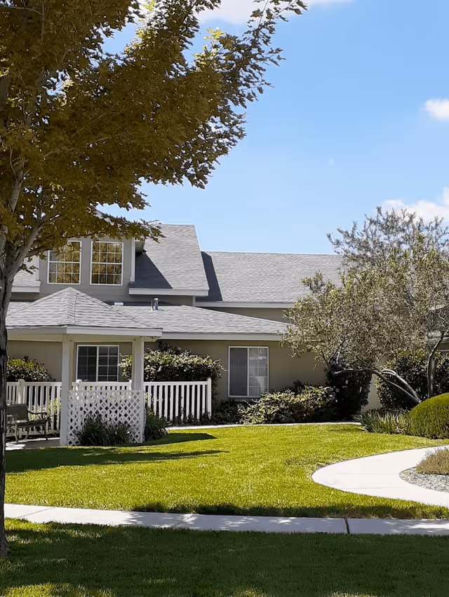 A sunny outdoor view of a senior living facility with a well-maintained green lawn, a white gazebo with lattice sides, trees, shrubs, and a light gray building with multiple windows in the background under a clear blue sky.
