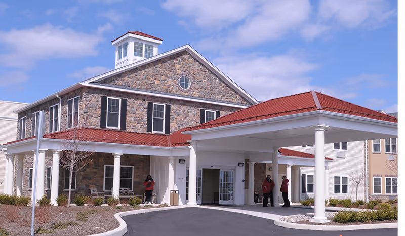 Exterior view of a senior living facility named The Birches at Newtown, featuring a stone facade with black shutters, a red metal roof, white columns supporting a covered entrance, and a paved driveway. Three people are standing near the entrance under the covered area.