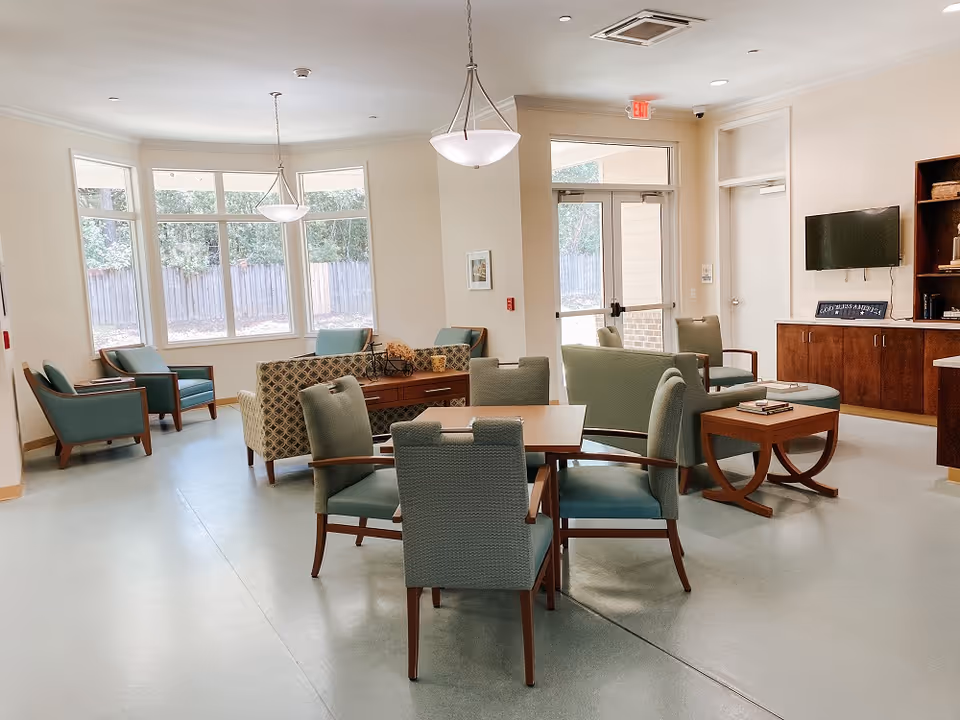 A bright and spacious assisted living community common area with multiple seating arrangements including armchairs, sofas, and tables. Large windows let in natural light, and there is a wall-mounted TV above a wooden cabinet. The room has light-colored walls and flooring, with pendant lights hanging from the ceiling.