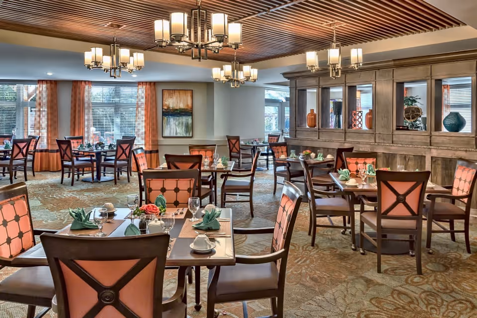 A well-lit dining room with multiple wooden tables and chairs featuring orange cushioned backs. Each table is set with folded green napkins, cups, glasses, and small floral centerpieces. The room has large windows with orange curtains, decorative ceiling lights, and a wooden cabinet with vases and decor items.