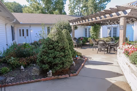 Sunny courtyard with a wooden pergola, outdoor table and chairs, and landscaped garden beds beside a single-story building.