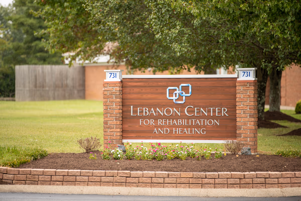 Outdoor view of a brick and wood sign for Lebanon Center for Rehabilitation and Healing, surrounded by a small flower bed and greenery with trees and a building in the background.