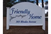 White freestanding outdoor sign that reads "Friendly Home" with "303 Rhodes Avenue" beneath it, set among trees.