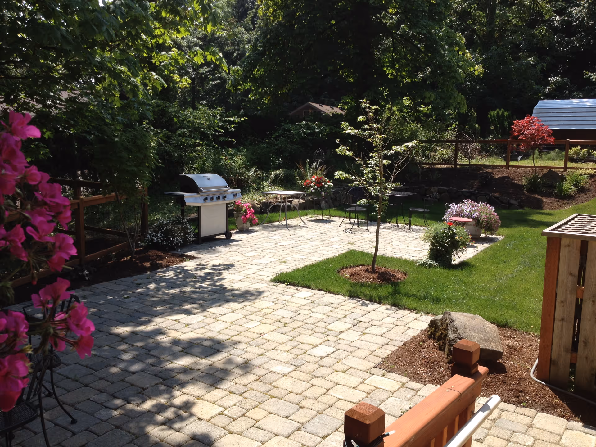 A sunny outdoor patio area with a stone-paved floor, a barbecue grill, several metal chairs and tables, surrounded by green grass, trees, and colorful flowers. There is a wooden fence and a small shed in the background.
