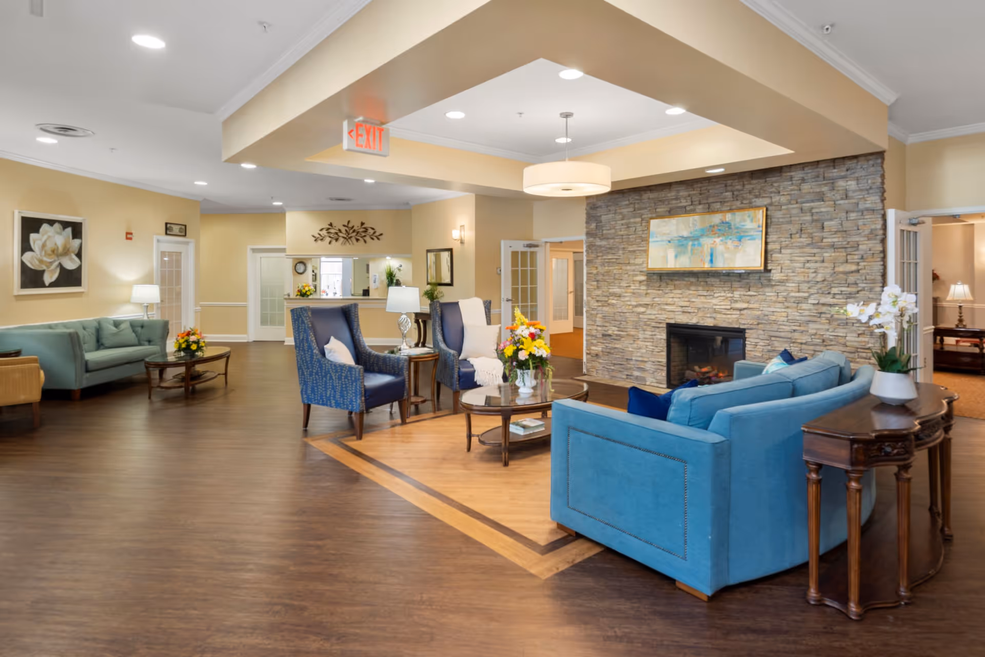 A spacious and well-lit senior living facility common area with comfortable seating including blue sofas and armchairs arranged around a coffee table with a flower arrangement. The room features a stone accent wall with a fireplace and a framed artwork above it. The floor is a combination of wood and laminate, and there are decorative lamps and plants placed around the room. Doors and an exit sign are visible in the background.