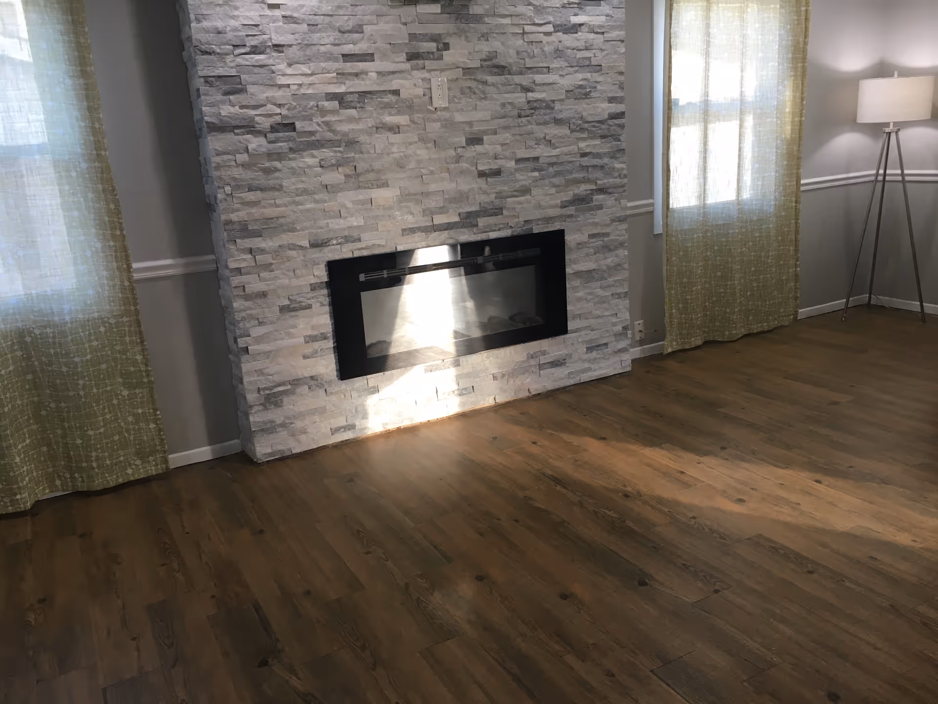 A room with wooden flooring, a modern stone fireplace in the center of the wall, two windows with light green curtains on either side of the fireplace, and a tall floor lamp with a white shade in the corner.