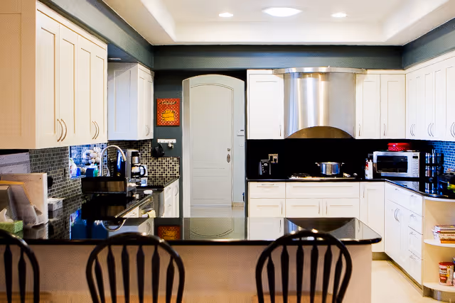 Modern kitchen with white cabinets, black countertops, a stainless steel range hood, microwave, coffee maker, and a kitchen island with three black chairs in the foreground.