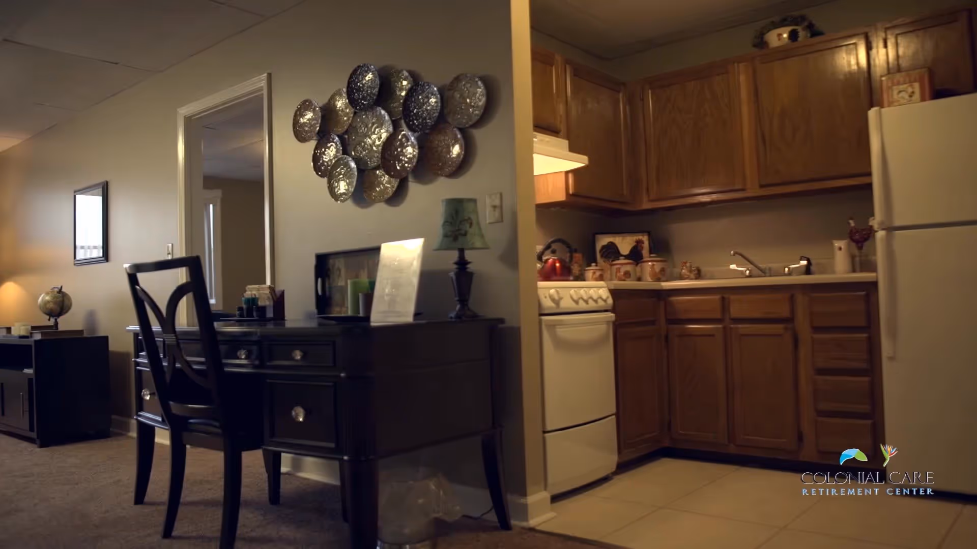 Interior view of a retirement center apartment showing a small kitchen with wooden cabinets, a white stove, and a refrigerator. Adjacent to the kitchen is a dark wooden desk with a chair, a lamp, and decorative items on the wall above it. The room has beige walls and carpeted flooring in the living area.