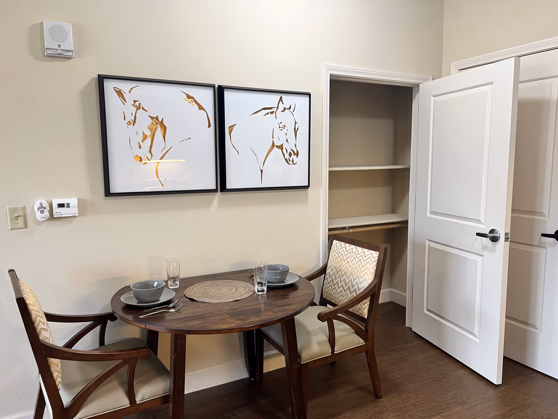 Small dining nook with a round wooden table set for two, two upholstered chairs, framed horse artwork on the wall, and an open closet door.