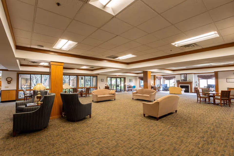 Spacious assisted living facility common area with multiple seating arrangements including beige sofas and dark armchairs, wooden columns, carpeted floor, large windows letting in natural light, and a fireplace in the background.