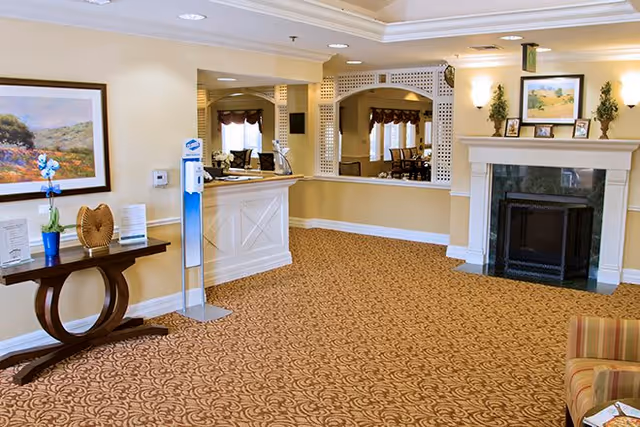 A spacious and well-lit common area in a senior living facility featuring a patterned carpet, a white reception desk, a fireplace with decorative items on the mantle, a small table with a blue vase and a plant, and framed artwork on the walls. There are also windows with curtains and a seating area partially visible.