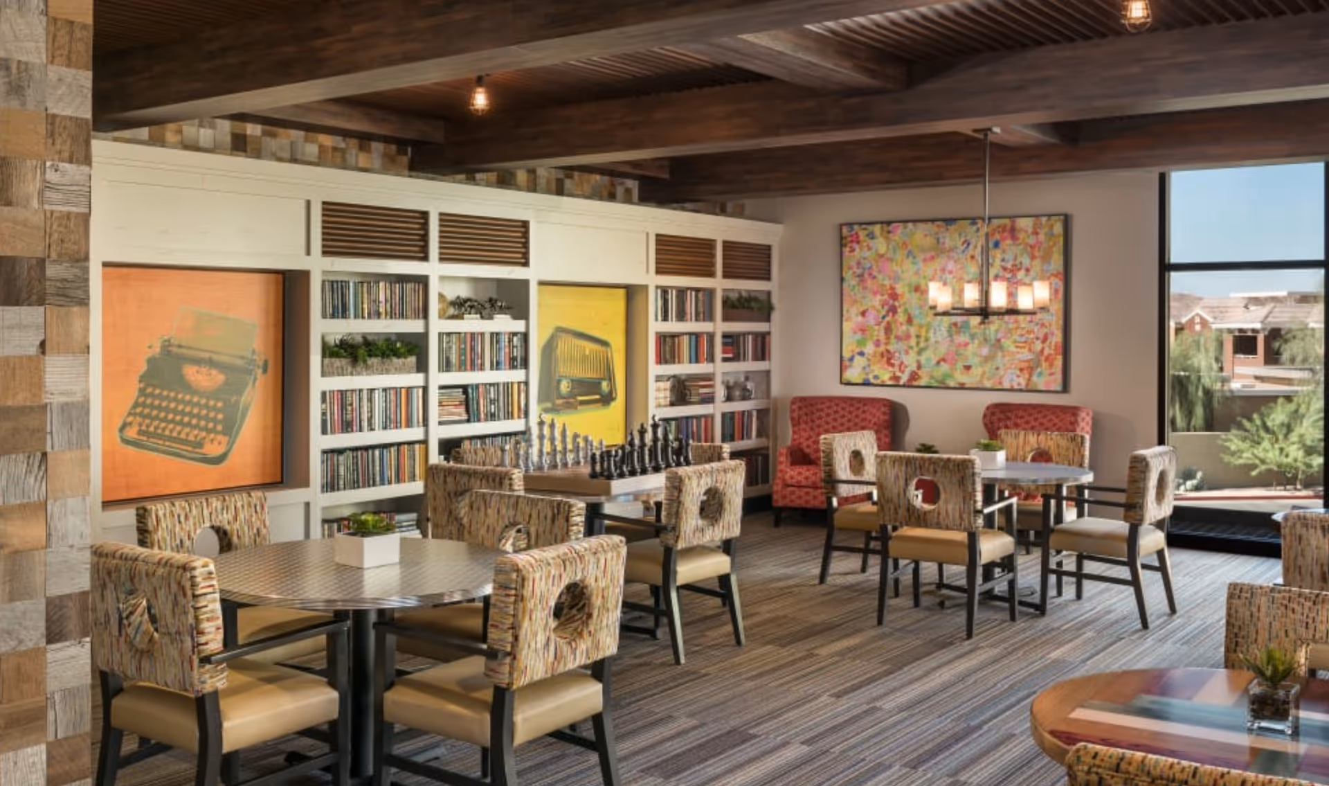 Communal lounge with tables and patterned chairs, bookshelves, colorful artwork, and a chess set near a large window.