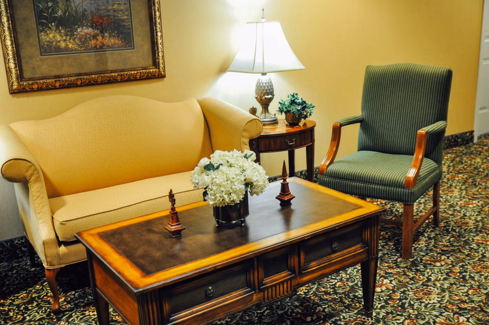 A cozy sitting area with a yellow upholstered sofa, a green striped armchair, and a wooden coffee table with decorative items and a vase of white flowers. A side table with a lamp and a small plant is placed next to the sofa, and a framed floral painting hangs on the wall above.