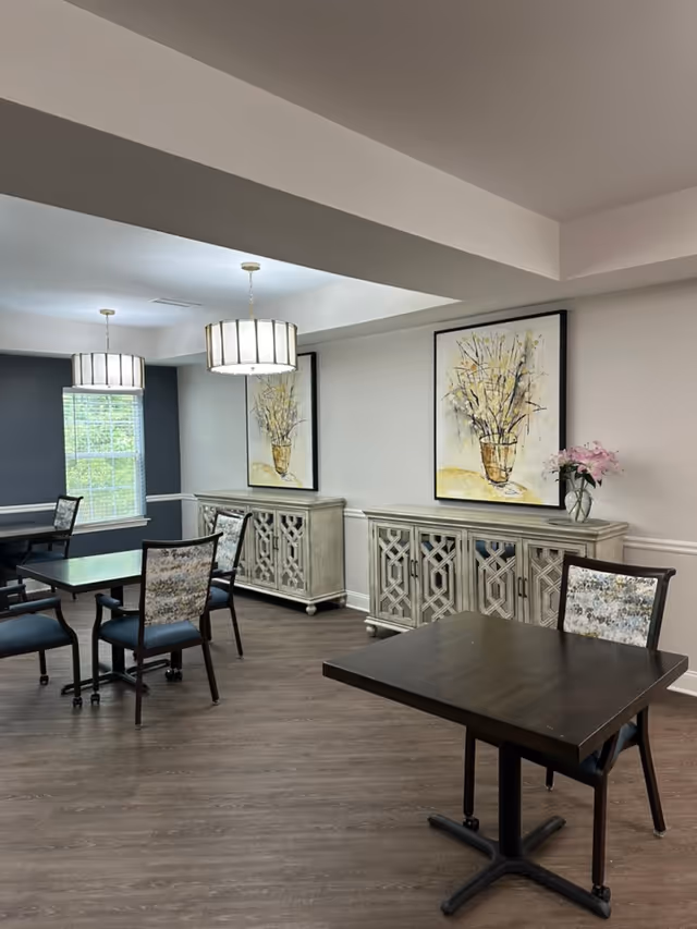 A dining area with dark wooden tables and chairs featuring patterned upholstery. The room has light-colored walls with two large framed paintings of floral arrangements. Two pendant lights hang from the ceiling, and there is a window letting in natural light. Two decorative sideboards with intricate designs are placed against the wall, one adorned with a vase of pink flowers.