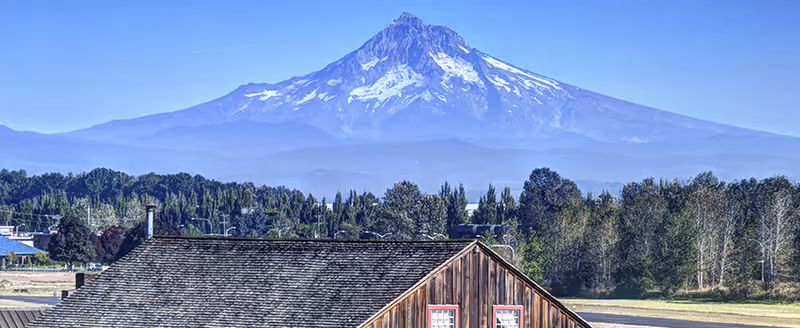 A rustic wooden building with a shingled roof in the foreground, surrounded by trees and greenery, with a large snow-capped mountain in the background under a clear blue sky.