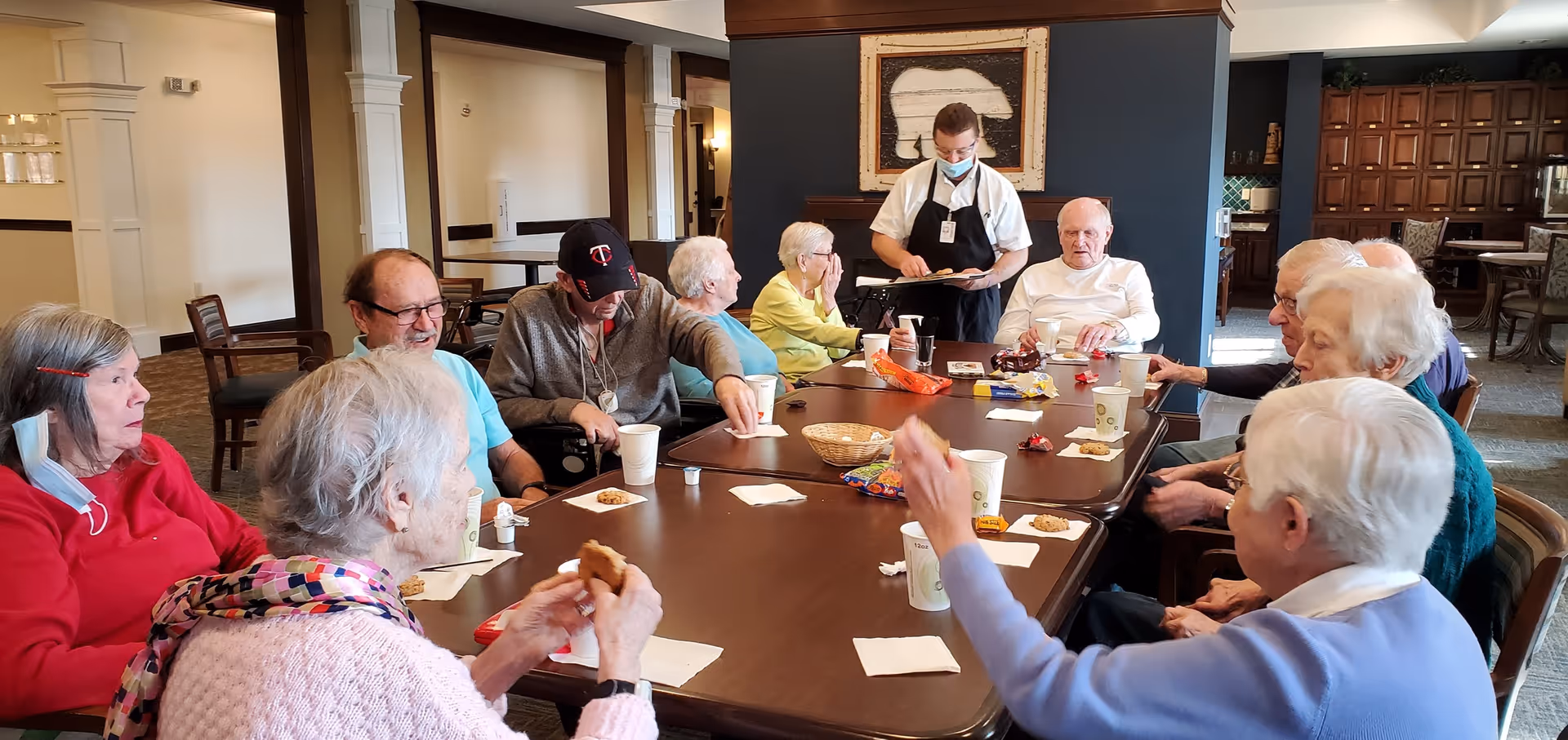 A group of elderly residents seated around a long table eating snacks while a masked staff member serves them in a communal dining/activity room.