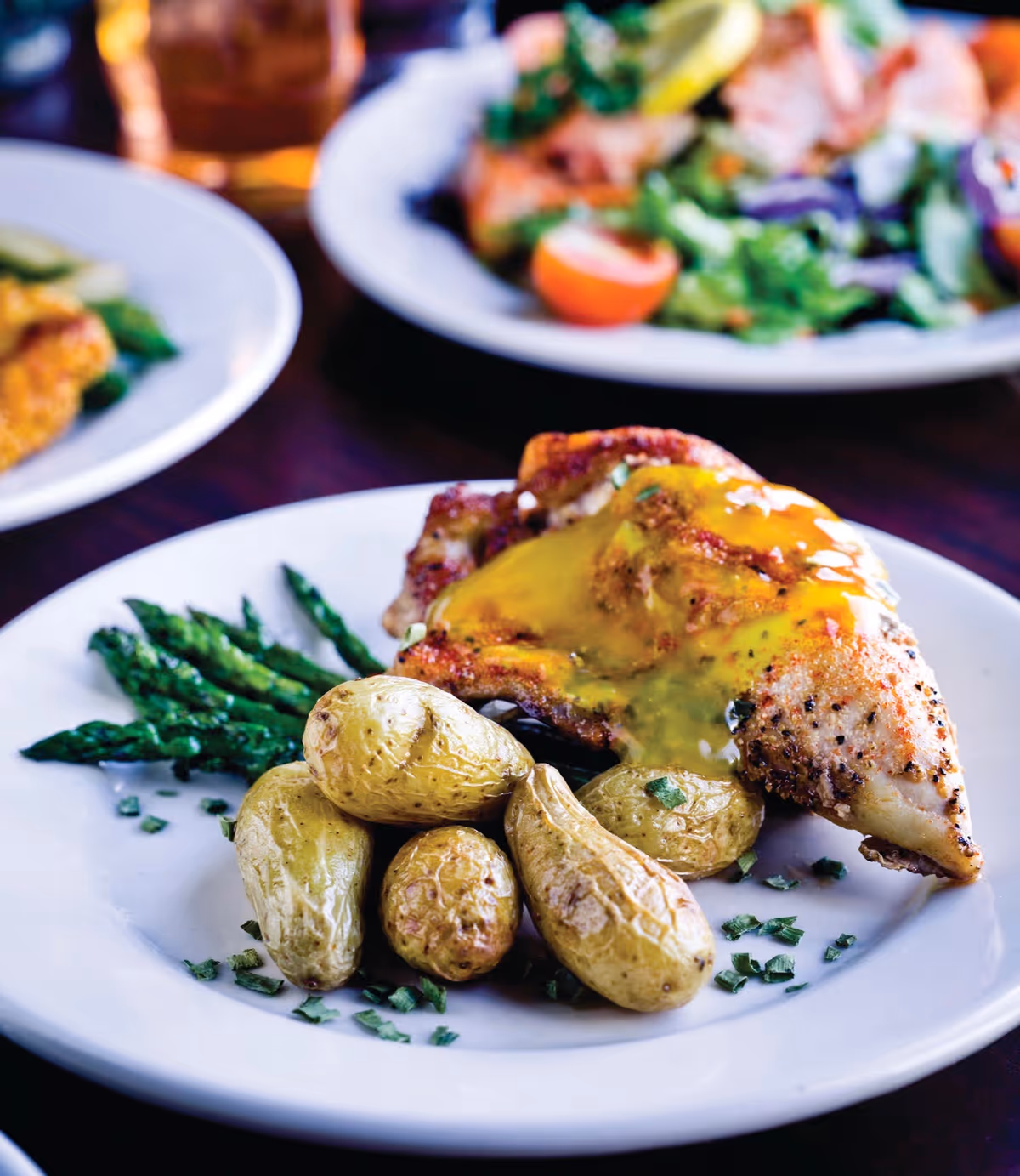 A plated meal featuring roasted fingerling potatoes, grilled asparagus, and a piece of grilled chicken breast topped with a yellow sauce, with blurred plates of food in the background.