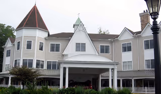 Exterior view of a multi-story senior living facility with beige siding, white trim, and a prominent peaked roof structure above the entrance. There is greenery and a lamppost in the foreground.