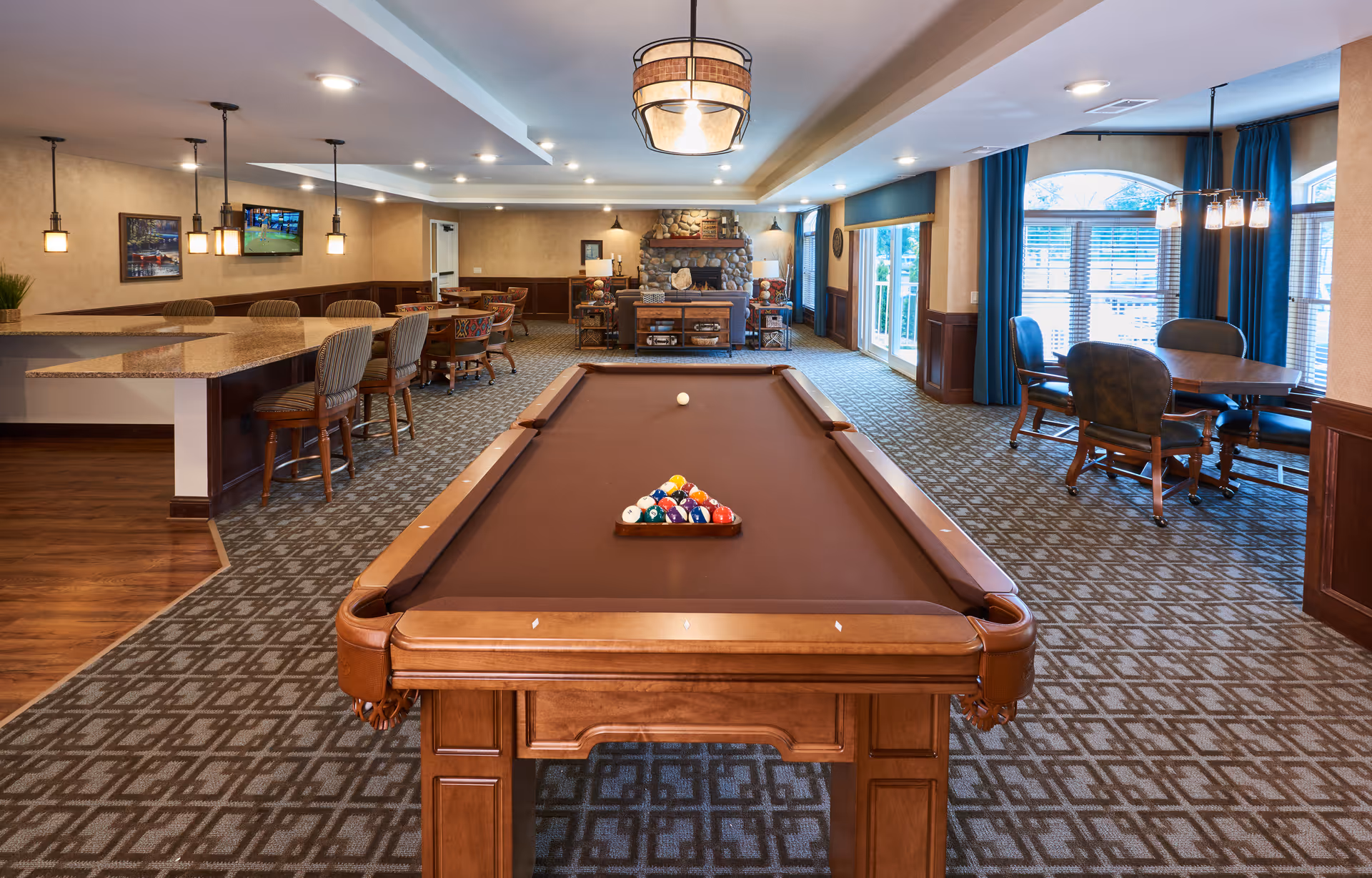 A spacious recreational room featuring a pool table with balls arranged for a game, surrounded by a patterned carpet. The room includes a seating area with tables and chairs near large windows with blue curtains, a stone fireplace in the background, and a counter with bar stools on the left side. The room is well-lit with ceiling lights and pendant lamps.