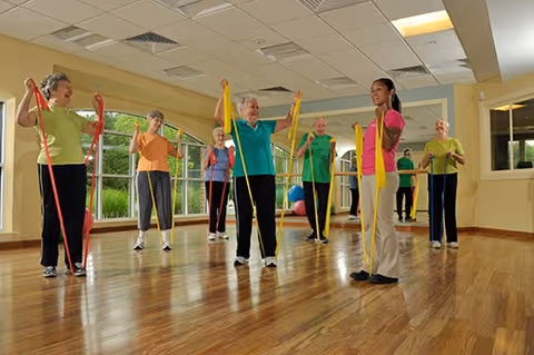 A group of elderly individuals participating in a fitness class indoors, each holding colorful resistance bands while following the instructor's guidance in a spacious room with wooden floors and large windows.